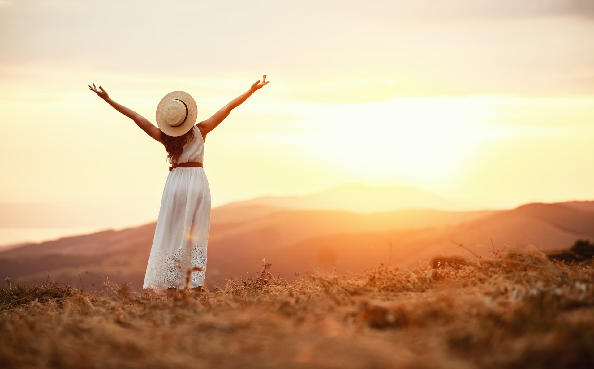 A woman in a white dress standing on a hill at sunset with arms raised, feeling free and renewed