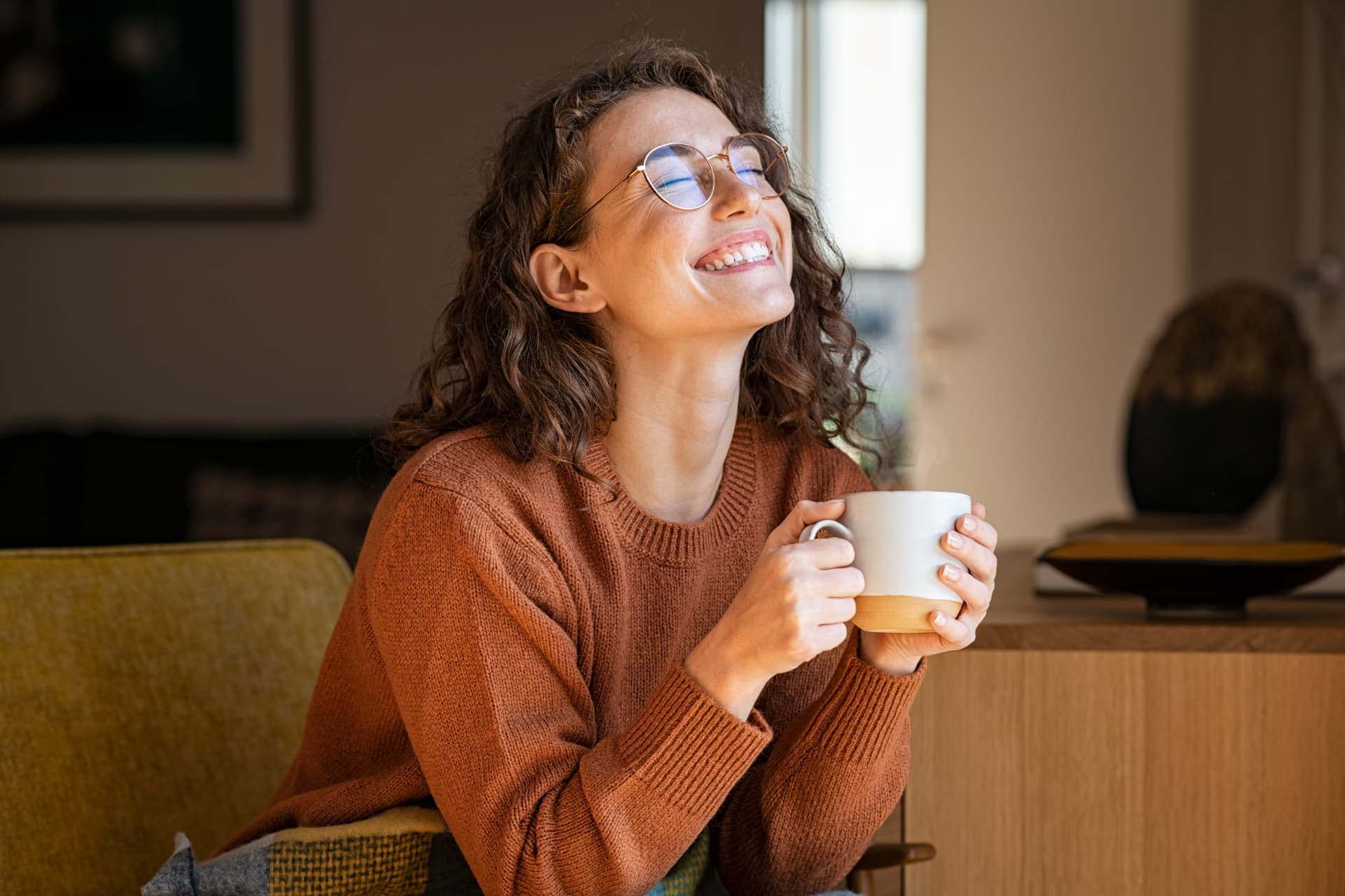 A smiling woman in a cozy rust sweater relaxing with a warm mug, looking peaceful and content